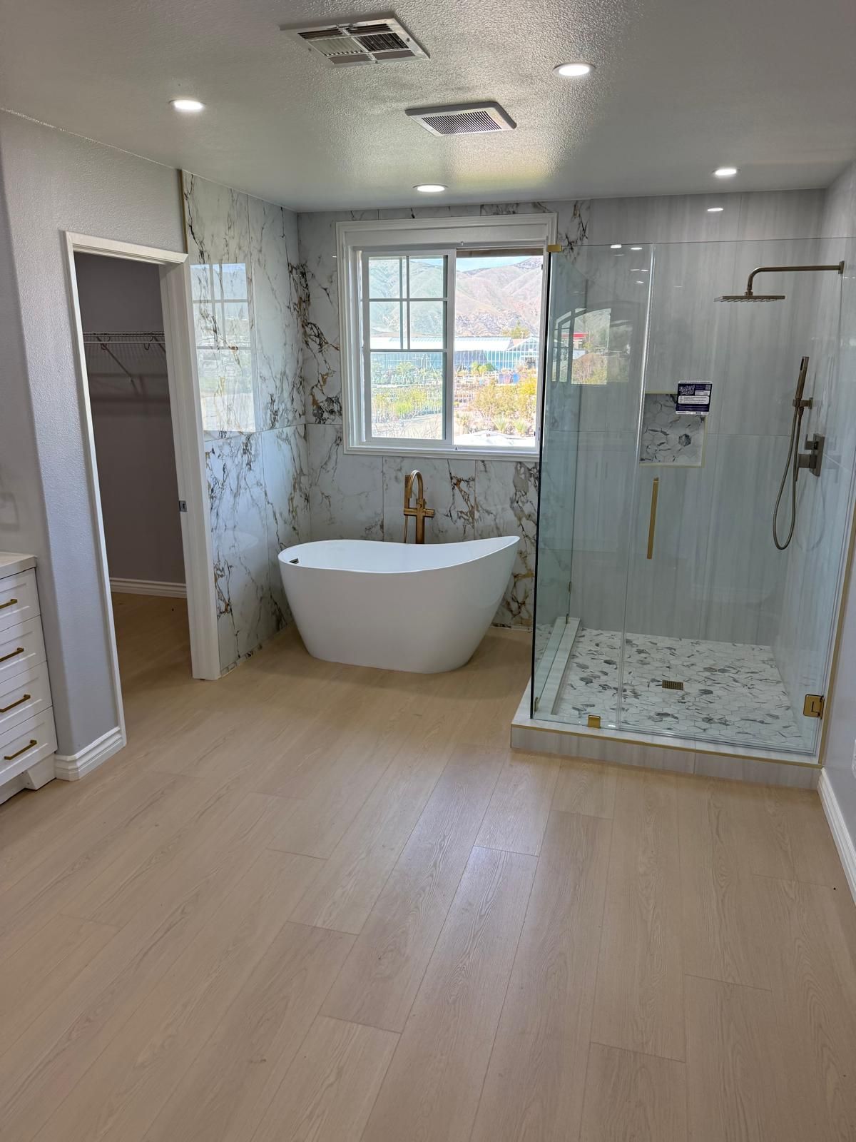 The same bathroom rebuilt with a freestanding tub under the window, a glass walk-in shower alongside, marble-look tile, and wide plank wood-look flooring.
