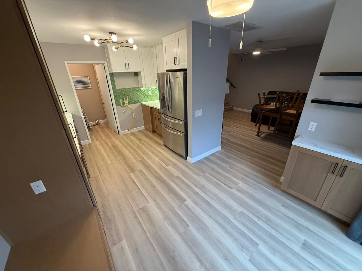 Open plan kitchen with white upper cabinets, a green patterned tile backsplash, a stainless refrigerator, and wide plank floors running to the dining area.