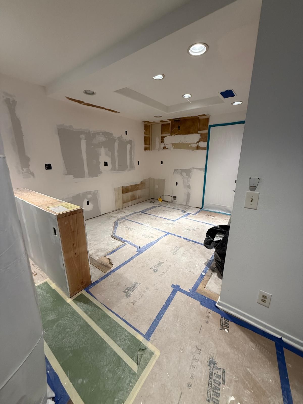 Kitchen with drywall patched and taped, recessed lights in the ceiling, a first cabinet box set in place, and the subfloor covered in taped protection board.